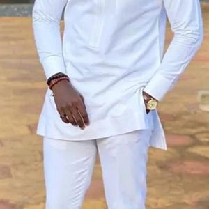 May include: A white, long-sleeved shirt and matching pants. The shirt has a mandarin collar and a pocket with a gold-colored design. A person is wearing a watch and a bracelet. The outfit is a traditional style.