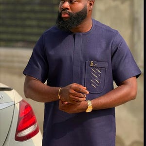 May include: Navy blue short-sleeved tunic with a pocket featuring a gold design. The tunic has a mandarin collar and a relaxed fit. A gold watch and bracelet are visible. The outfit is suitable for formal or casual wear.