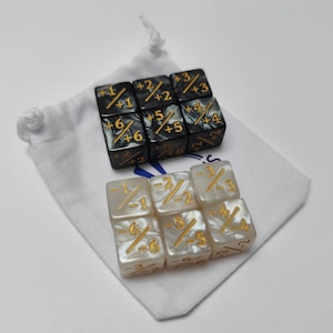 Op de afbeelding: Een set van twaalf dobbelstenen, zes zwarte en zes witte, met gouden markeringen. De zwarte dobbelstenen hebben een marmer effect en tonen positieve en negatieve getallen. De witte dobbelstenen hebben een parelmoer afwerking en tonen breuken. De dobbelstenen worden gepresenteerd met een wit trekkoordzakje.