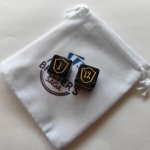 May include: Two black dice with gold numbering and shield designs, resting on a white drawstring bag. The dice are marked with the numbers 1 and 12. The bag has the text "BARRER'S SAGA" printed on it.