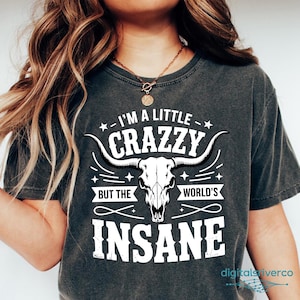 May include: Dark gray t-shirt with the text "I'M A LITTLE CRAZZY BUT THE WORLD'S INSANE" and a white bull skull graphic. The shirt is a crew neck and has short sleeves.