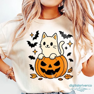 May include: Cream-colored Halloween-themed t-shirt featuring a cartoon cat sitting on a carved orange pumpkin. Black bats, stars, and leaves surround the cat and pumpkin design. The shirt has a crew neck and short sleeves.