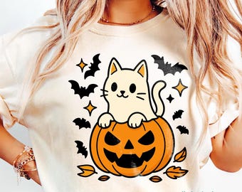 May include: Cream-colored Halloween-themed t-shirt featuring a cartoon cat sitting on a carved orange pumpkin. Black bats, stars, and leaves surround the cat and pumpkin design. The shirt has a crew neck and short sleeves.