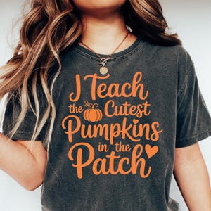 Könnte beinhalten: Dunkelgraues T-Shirt mit dem Text "I Teach the Cutest Pumpkins in the Patch" in Orange. Das Shirt zeigt eine Kürbisgrafik und ein Herz. Das Shirt ist ein bequemer, lässiger Stil.