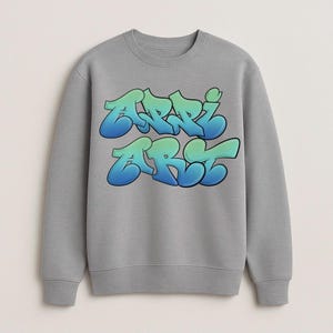 May include: A gray sweatshirt with the word "ARRI ART" in a graffiti-style font. The text is gradient, transitioning from blue to green. The sweatshirt has a crew neck and long sleeves.