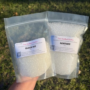 May include: Two clear plastic bags filled with white aroma beads. One bag is labeled "BLACK ICE" and the other "LEATHER." The labels include the Etsy shop name and website address. The background is blurred green grass.