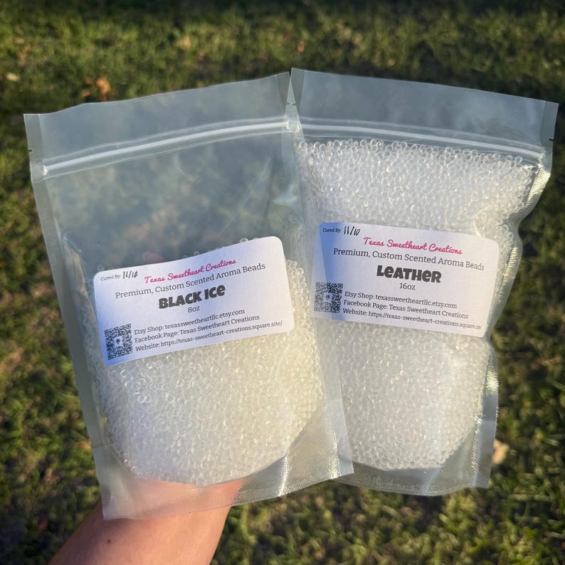 May include: Two clear plastic bags filled with white aroma beads. One bag is labeled "BLACK ICE" and the other "LEATHER." The labels include the Etsy shop name and website address. The background is blurred green grass.