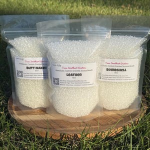 May include: Three clear plastic bags filled with white aroma beads, each labeled with a different scent: "Butt Naked", "Leather", and "Bombshell". The bags are displayed on a wooden slice, with green grass in the background.