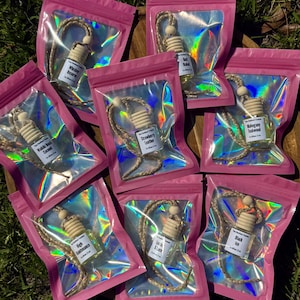 May include: A selection of car air fresheners in small glass bottles with wooden tops, displayed in pink holographic pouches. Each bottle is labelled with a scent name, such as "Strawberry Leather" and "Mahogany Teakwood". The bottles are attached to jute cords.
