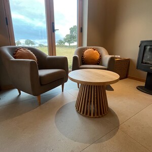 May include: Two grey armchairs with orange cushions and a round wooden coffee table. The table has a slatted base. A wicker basket and a fireplace are also visible in the room.