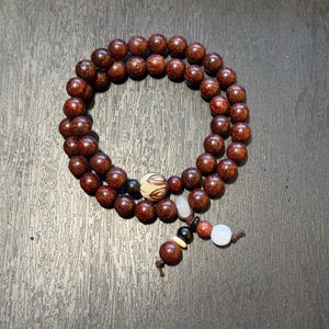 May include: A circular beaded bracelet composed of numerous dark brown wooden beads. The bracelet features a single patterned bead with brown and cream swirls, two smooth black beads, two translucent white beads, and one small tan bead.