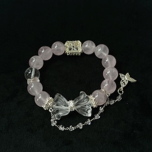 Könnte beinhalten: Ein Armband aus Rosenquarzperlen mit einer durchsichtigen Kristallschleife und einem silbernen Schmetterlingsanhänger. Das Armband hat einen silbernen Verschluss und kleine Kristallakzente. Die Perlen sind zartrosa.