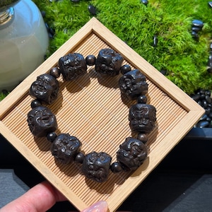 Peut inclure: Un bracelet en perles de bois brun foncé avec des visages sculptés sur chaque grosse perle, séparées par de plus petites perles d'espacement rondes et sombres. Le bracelet est disposé en cercle sur un plateau carré à lattes en bambou.
