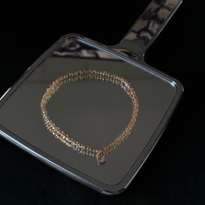 Puede incluir: Un collar dorado con un pequeño colgante, exhibido en un espejo de mano plateado. El collar está hecho de pequeñas cuentas iridiscentes. El espejo refleja el collar sobre un fondo negro.