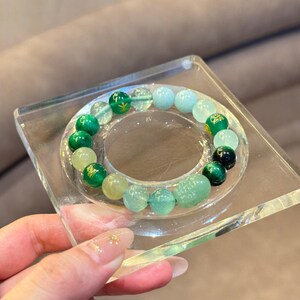Może przedstawiać: Bransoletka z mieszanką zielonych, jasnoniebieskich i przezroczystych koralików. Bransoletka jest prezentowana na przezroczystym, kwadratowym stojaku akrylowym. Koraliki różnią się rozmiarem i odcieniem, tworząc kolorowy i teksturowany dodatek.