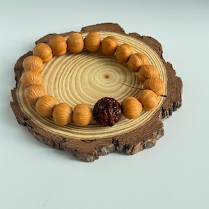 May include: A beaded bracelet featuring light brown, walnut-shaped beads and a single, dark red bead. The bracelet is displayed on a small, round wooden slice with a natural bark edge, set against a white background.