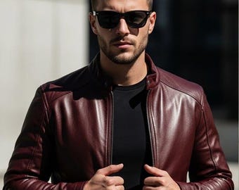 Puede incluir: Chaqueta de cuero burdeos con cremallera y camiseta negra debajo. La chaqueta tiene cuello alto y se lleva con gafas de sol negras. La persona mira directamente a la cámara.