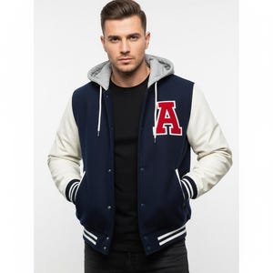 Puede incluir: Chaqueta universitaria azul marino con mangas blancas y forro de capucha gris. La chaqueta presenta una gran letra "A" roja en el pecho, rayas blancas en los puños y el dobladillo, y una parte delantera abotonada. Se lleva sobre una camiseta negra.