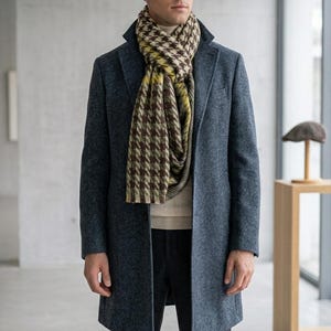 May include: A charcoal blue wool-blend overcoat worn over a cream-colored turtleneck sweater and dark gray trousers. A long scarf featuring a brown, cream, and yellow houndstooth pattern is wrapped around the neck, resting over the coat lapels.