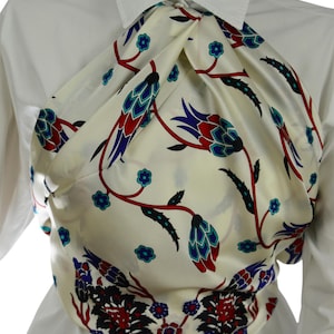 Puede incluir: Bufanda de seda color crema con un estampado floral en rojo, azul y turquesa. La bufanda está drapeada sobre una camisa blanca con cuello. El diseño floral presenta flores y hojas estilizadas.