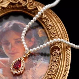 Puede incluir: Un collar de perlas con un colgante de oro con una gran gema roja en forma de lágrima. El colgante está rodeado de perlas más pequeñas y piedras transparentes. El collar se exhibe frente a un cuadro con marco dorado.