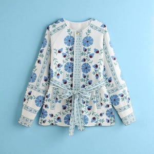 Könnte beinhalten: Weißes Jackett mit blauem Blumenmuster und passendem Bindegürtel. Die Jacke hat lange Ärmel und einen runden Ausschnitt. Das florale Design umfasst blaue Blumen und rote Akzente. Die Jacke hat einen Knopfverschluss.