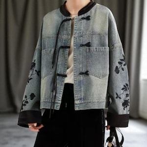 May include: A light blue denim jacket with black floral embroidery on the sleeves. The jacket has a mandarin collar, black frog closures, and black trim on the collar and cuffs. It features two front pockets.