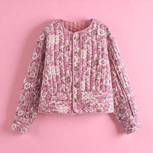 May include: A quilted jacket in shades of pink and white, featuring a floral pattern. The jacket has a round neckline, long sleeves, and gold-colored buttons. The background is a solid pink.