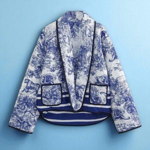 Puede incluir: Chaqueta acolchada azul y blanca con un estampado de toile de Jouy con tigres y otros animales. La chaqueta tiene cuello chal, dos bolsillos y ribete negro. El bajo tiene un diseño de rayas azules y blancas.