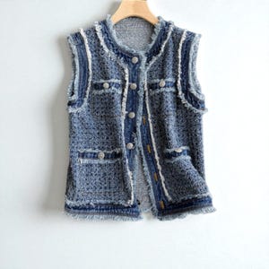 May include: A sleeveless denim vest with a tweed-like texture. The vest has a frayed edge design, two front pockets, and silver buttons. It hangs on a wooden hanger against a white background.