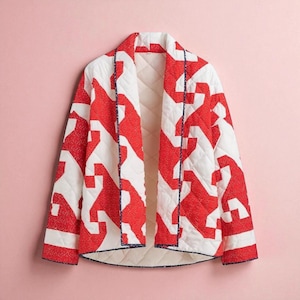 May include: A quilted jacket with a geometric red and white pattern. The jacket features a shawl collar and a quilted lining. It is trimmed with navy blue binding. The jacket is shown against a pink backdrop.