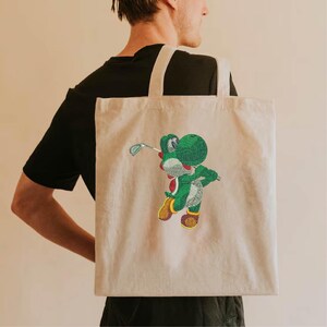 Puede incluir: Bolso tote beige con un personaje Yoshi verde bordado jugando al golf. El bolso tiene forma rectangular y una correa para el hombro. El diseño de Yoshi incluye detalles en rojo, blanco, amarillo y marrón. El bolso se lleva sobre el hombro.