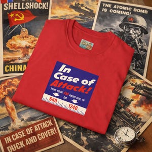 May include: A red t-shirt with the text "In Case of Attack!" in a blue and white design. The shirt also features a radio dial graphic. The shirt is surrounded by vintage propaganda posters and a pocket watch.