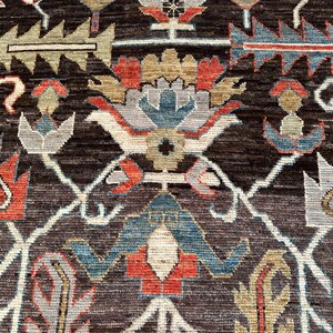 K&ouml;nnte beinhalten: Ein dunkelbrauner Teppich mit einem sich wiederholenden Muster aus stilisierten Blumen- und geometrischen Motiven in Rot-, Blau-, Hellgrau- und Beiget&ouml;nen. Das Design umfasst miteinander verbundene wei&szlig;e Linien, die ein rautenf&ouml;rmiges Gitter bilden.