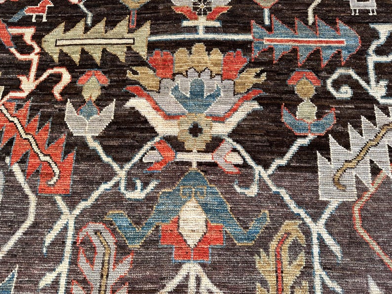 K&ouml;nnte beinhalten: Ein dunkelbrauner Teppich mit einem sich wiederholenden Muster aus stilisierten Blumen- und geometrischen Motiven in Rot-, Blau-, Hellgrau- und Beiget&ouml;nen. Das Design umfasst miteinander verbundene wei&szlig;e Linien, die ein rautenf&ouml;rmiges Gitter bilden.