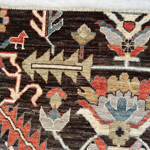 K&ouml;nnte beinhalten: Ein dunkelbrauner Teppich mit einem sich wiederholenden Muster aus stilisierten Blumenmotiven, geometrischen Formen und kleinen Tierfiguren in Rot, Hellblau, Beige und Grau. Der Teppich hat eine sichtbare Webstruktur und einen Fransenrand.