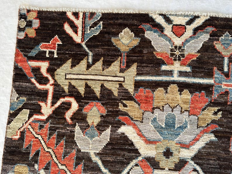 K&ouml;nnte beinhalten: Ein dunkelbrauner Teppich mit einem sich wiederholenden Muster aus stilisierten Blumenmotiven, geometrischen Formen und kleinen Tierfiguren in Rot, Hellblau, Beige und Grau. Der Teppich hat eine sichtbare Webstruktur und einen Fransenrand.