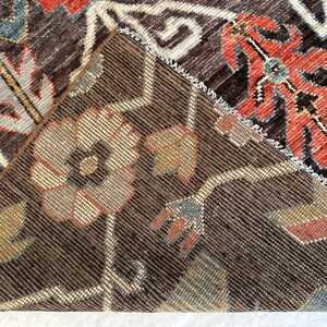 K&ouml;nnte beinhalten: Ein dunkelbrauner Teppich mit einer umgeschlagenen Ecke, die die kontrastierende Unterseite zeigt. Der Teppich weist ein florales Muster mit komplexen Motiven in Rot-, Blau-, Beige- und Wei&szlig;t&ouml;nen auf und zeigt eine dichte Webstruktur.