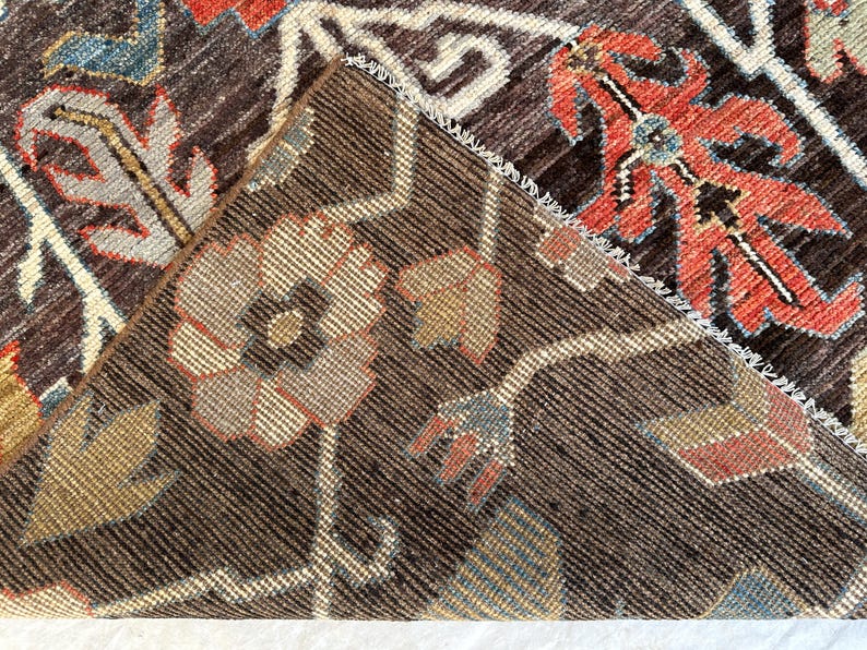 K&ouml;nnte beinhalten: Ein dunkelbrauner Teppich mit einer umgeschlagenen Ecke, die die kontrastierende Unterseite zeigt. Der Teppich weist ein florales Muster mit komplexen Motiven in Rot-, Blau-, Beige- und Wei&szlig;t&ouml;nen auf und zeigt eine dichte Webstruktur.