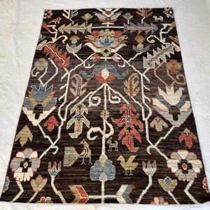 K&ouml;nnte beinhalten: Ein rechteckiger Teppich mit dunkelbraunem Hintergrund und einem symmetrischen Muster aus stilisierten Blumen, Bl&auml;ttern und kleinen Vogelmotiven in Rot-, Blau-, Beige- und Graut&ouml;nen, angeordnet in einem sich wiederholenden botanischen Design.