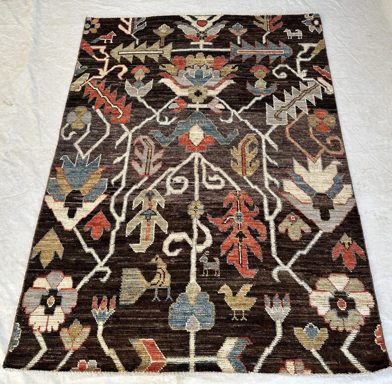 K&ouml;nnte beinhalten: Ein rechteckiger Teppich mit dunkelbraunem Hintergrund und einem symmetrischen Muster aus stilisierten Blumen, Bl&auml;ttern und kleinen Vogelmotiven in Rot-, Blau-, Beige- und Graut&ouml;nen, angeordnet in einem sich wiederholenden botanischen Design.