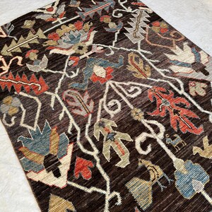 K&ouml;nnte beinhalten: Ein rechteckiger Teppich mit dunkelbraunem Hintergrund und einem sich wiederholenden Muster aus stilisierten Blumenmotiven und Vogelfiguren in Rot-, Blau-, Beige- und Graut&ouml;nen, angeordnet in einem symmetrischen, rankenartigen Design.