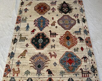 Könnte beinhalten: Ein rechteckiger Teppich mit cremefarbener Basis, der ein wiederkehrendes Muster aus bunten geometrischen Medaillons, stilisierten Tierfiguren und menschenähnlichen Formen in Blau-, Rot-, Braun- und Violetttönen mit Fransenrand zeigt.