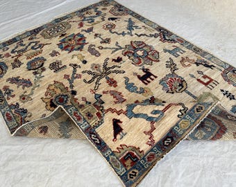 Könnte beinhalten: Ein rechteckiger Teppich mit cremefarbenem Grund und einem mehrfarbigen Muster aus stilisierten Blumenmotiven, geometrischen Formen und Tierfiguren in Blau-, Rot- und Brauntönen. Eine Ecke des Teppichs ist umgeschlagen und zeigt die Unterseite.