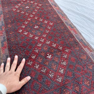 May include: A rectangular, hand-woven rug with a geometric pattern. The rug features a deep red central field with a repeating diamond design in dark grey and white. The border is a mix of red, grey, and brown tones.