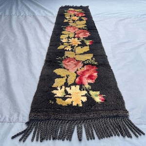 Puede incluir: Una alfombra larga y rectangular con un fondo marrón oscuro y un estampado floral. Las flores son principalmente rojas, amarillas y rosas, con hojas verdes. La alfombra tiene flecos en ambos extremos, agregando un elemento decorativo.