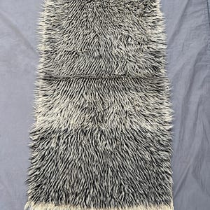 Könnte beinhalten: Ein rechteckiger Teppich mit zotteliger Textur und einem Muster aus schwarzen und cremefarbenen Fasern. Der Teppich hat einen Fransenbesatz an den kürzeren Kanten. Die langen Fasern erzeugen eine strukturierte Oberfläche. Der Teppich ist wahrscheinlich aus Wolle oder einem ähnlichen Material.