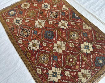 Könnte beinhalten: Ein rechteckiger Teppich mit tiefrotem Hintergrund und einem sich wiederholenden geometrischen Gittermuster. Das Design zeigt stilisierte Blumenmotive in Creme, Blau und Senfgelb, umrahmt von einer dunkelbraunen Bordüre mit Fransen an den Enden.