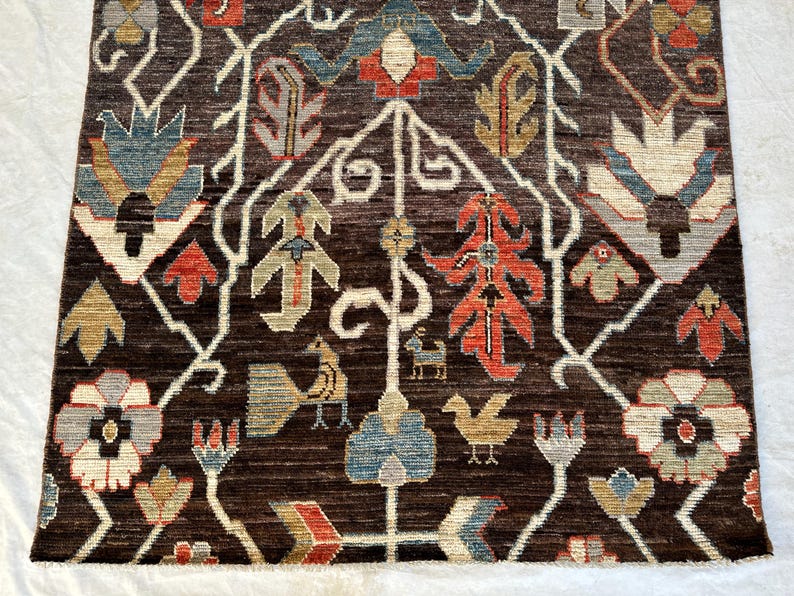 K&ouml;nnte beinhalten: Ein dunkelbrauner Teppich mit einem sich wiederholenden Muster aus stilisierten Blumenmotiven, Bl&auml;ttern und kleinen Vogelfiguren in Rot-, Blau-, Beige- und Cremet&ouml;nen. Das Design ist symmetrisch mit wei&szlig;en, rankenartigen Linien zwischen den Elementen.
