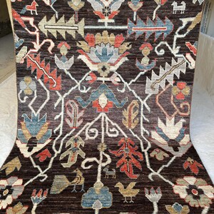 K&ouml;nnte beinhalten: Ein rechteckiger Teppich mit dunkelbraunem Hintergrund und einem symmetrischen Muster aus stilisierten Blumenmotiven, Bl&auml;ttern und kleinen Tierfiguren in Rot-, Blau-, Beige- und Wei&szlig;t&ouml;nen, abgeschlossen mit einer kurzen Franse am oberen Rand.